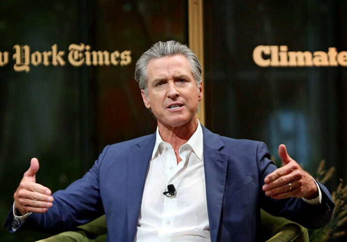 Gavin Newsom, wearing a blue sports jacket and white shirt gestures with both hands while speaking.