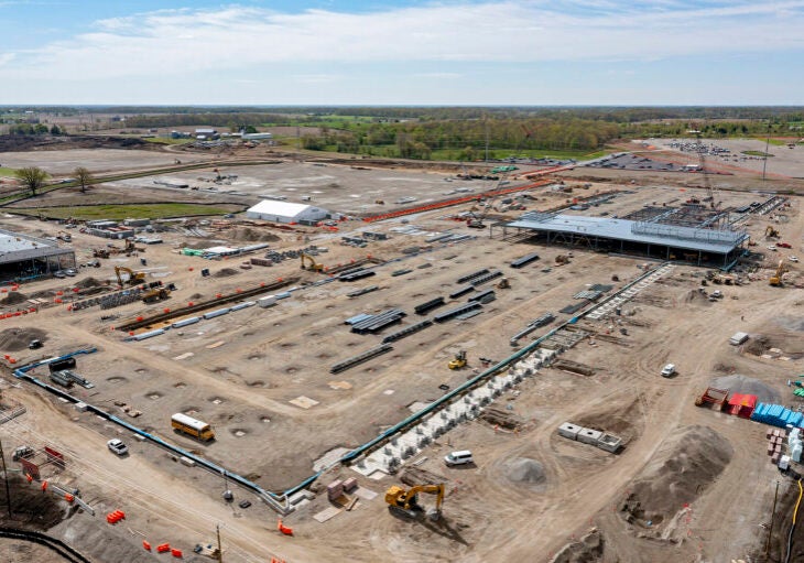 A large, graded, dirt construction site with machinery, vehicles and some buildings under construction, with flat farmland and trees visible in the distance.