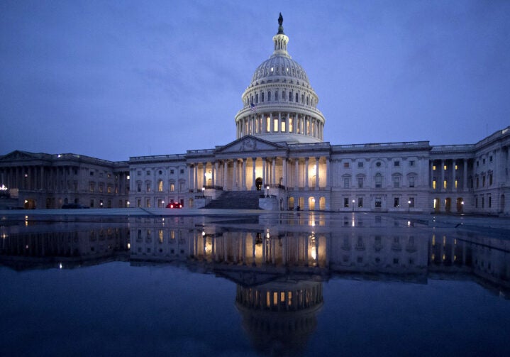 A wide photo of most of the U.S. Capitol building under a dark blue, cloudy sky. The building, with lights on, is reflected in water in the foreground.
