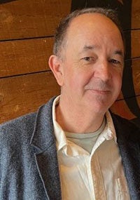 Geoff looks at the viewer with a slight smile. He is wearing a dark grey blazer, cream-colored collared, buttoned-down shirt, and light green undershirt. He's standing in front of a wood paneled wall.