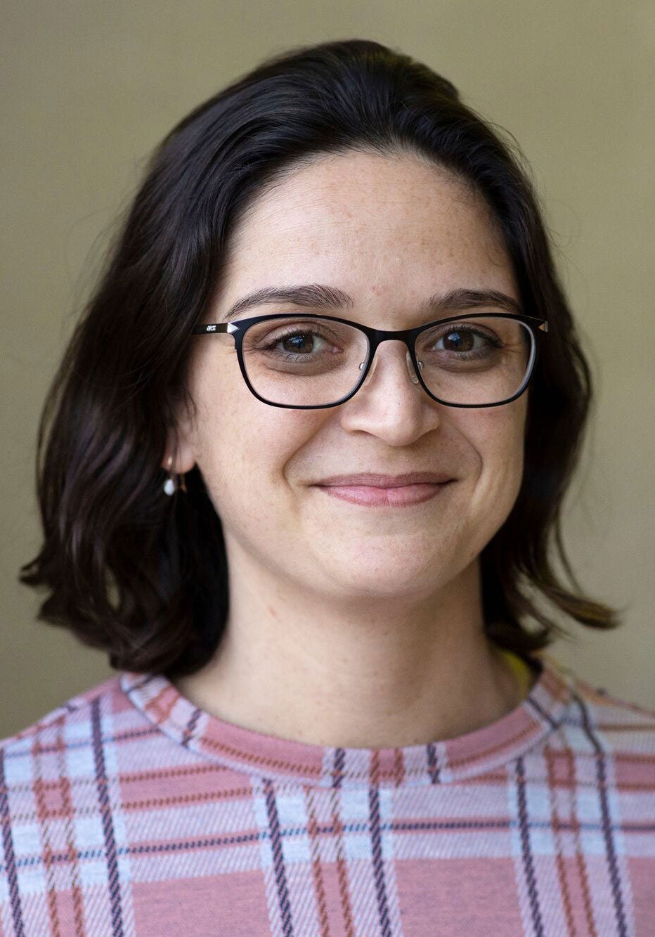 Gussie looks directly at the viewer with a slight smile. She's wearing a knit top with a coral, white, and dark blue flannel pattern. She's standing in front of a tan colored backdrop.