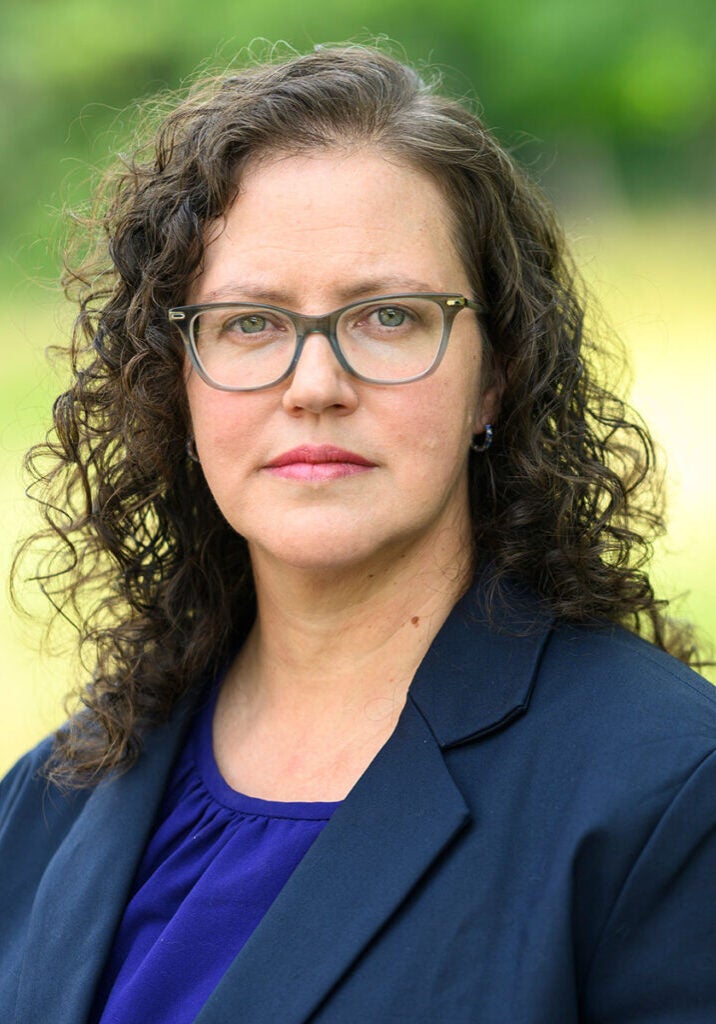 Hana Vizcarra, wearing grey glasses, a bright blue blouse, and a dark blue suit jacket, and standing in front of a soft focus light green background, looks directly at the viewer with a serious expression.