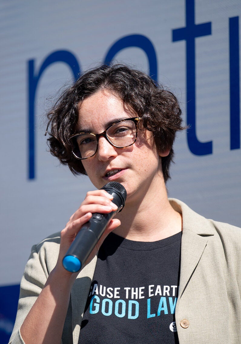 Hillary is speaking into a microphone in an outdoor setting; the sun is shining strongly on Hillary. Hillary is wearing a tan light jacket over a black t-shirt with the text, "Because the earth needs a good lawyer," printed across the front.