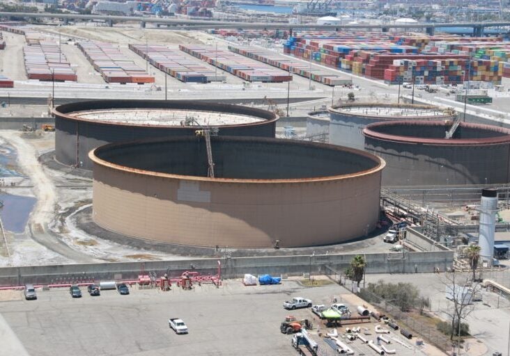 An aerial view of huge oil storage tanks. Two of the tanks in the foreground have large cranes in the middle of them. In the background are many stacked shipping containers in the Port of Long Beach, with ships and the water visible.