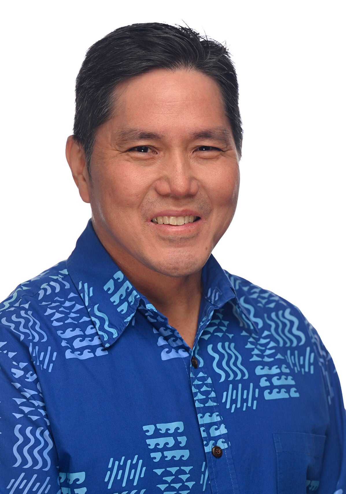 Isaac looks directly at the viewer with an open, friendly smile. He's wearing a bright blue collar buttoned-down shirt with light blue patterns of waves and water symbols. He is standing in front of a white backdrop.
