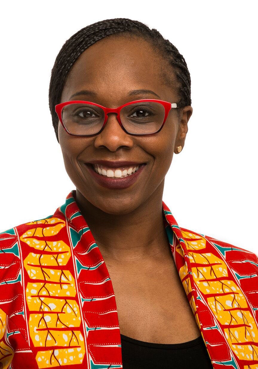 Jamila looks directly at the viewer with a friendly, open smile. She's wearing a brightly colored jacket with a red and yellow pattern and turquoise accents over a black top. She's standing in front of a white backdrop.