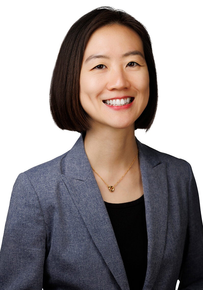 Jennifer looks directly at the viewer with a friendly, open smile. She's wearing a dark grey suit jacket over a black blouse. Jennifer's standing in front of a bright white backdrop.