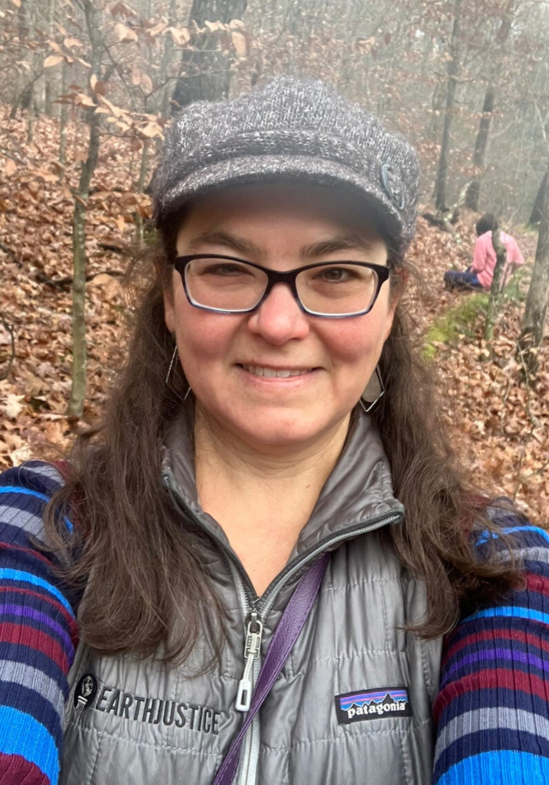Jenny looks directly at the viewer with a friendly open smile. She's outside in a forest, surrounded by a thick bed of fallen brown autumn leaves. A few leaves still cling to the tree branches. Jenny's wearing a black and white knitted cap, and a ribbed long sleeve shirt with stripes of bright blue, dark blue, grey, red, and purple colors, under a silver vest with the Earthjustice and Patagonia logo labels on either side of the front of the vest.