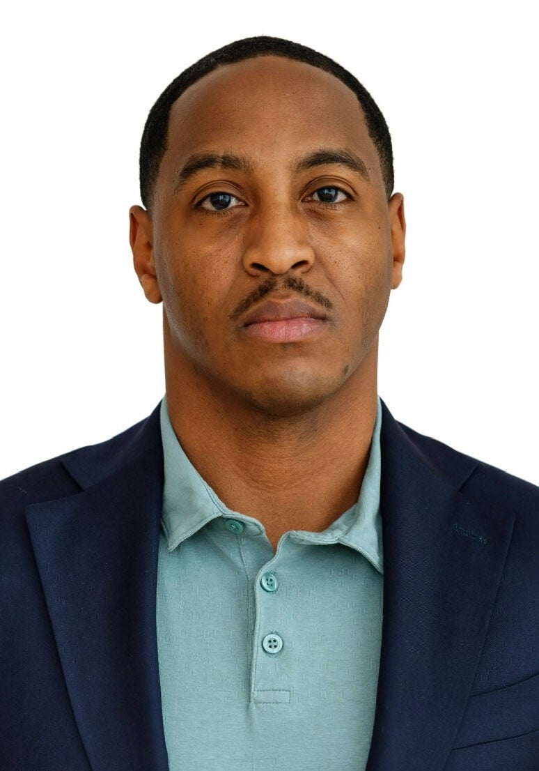 Jeremy looks directly at the viewer with a serious expression. He is wearing a dark blue suit jacket and a turquoise polo shirt. He is standing in front of a white backdrop.