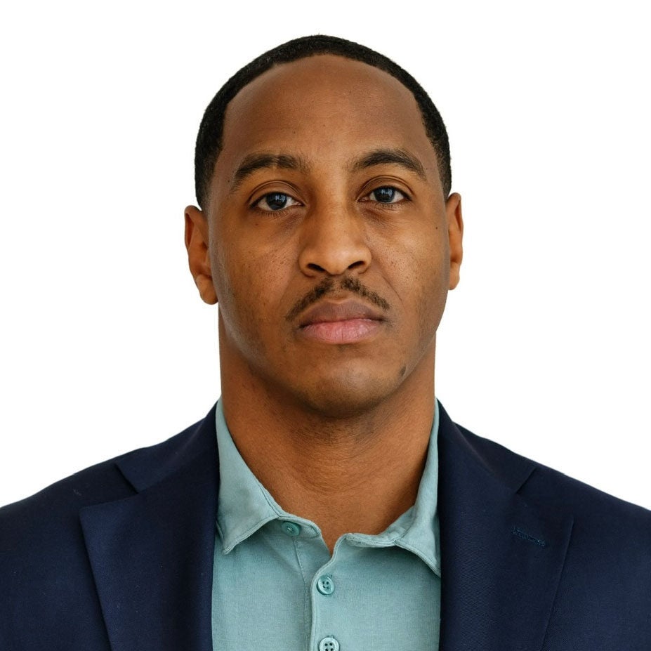 Jeremy looks directly at the viewer with a serious expression. He is wearing a dark blue suit jacket and a turquoise polo shirt. He is standing in front of a white backdrop.