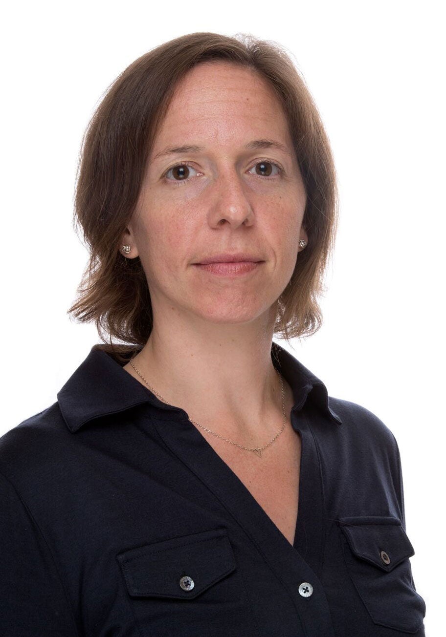 Jill looks directly at the viewer with a serious expression. She's wearing a collared, button-down black blouse, and stands against a white backdrop with her body slightly angled to the right.