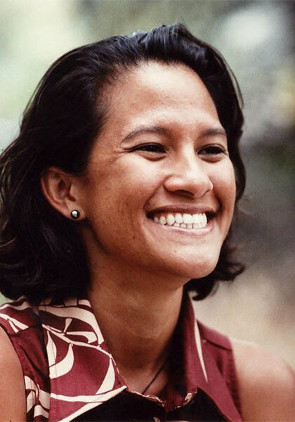 Kapua looks towards the viewer's right, with a broad, friendly smile. She's wearing a burgundy sleeveless top with a white floral pattern. The setting appears to be outdoors; the background is a blurred collage of dusky yellow and green colors.