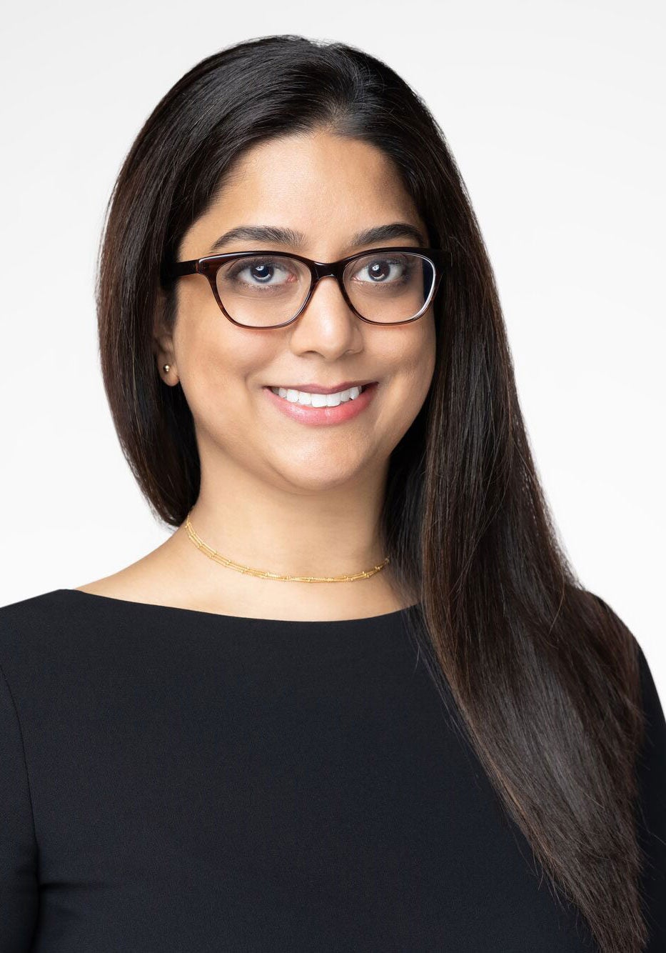 Kirti looks directly at the viewer, with a kind, open smile. She's wearing a black top and standing in front of a white backdrop.