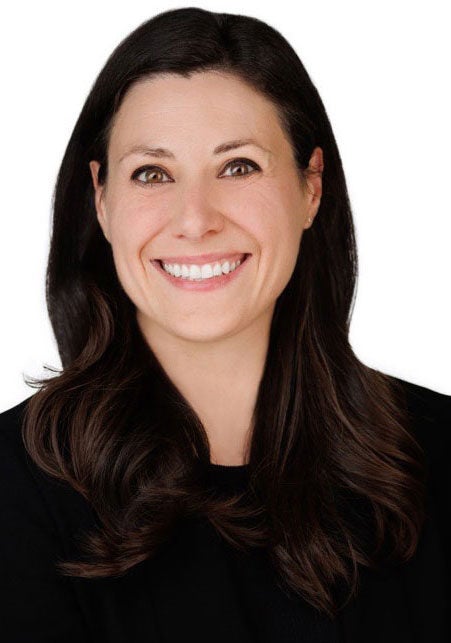Lauren looks at the viewer with a friendly, open smile. She's wearing a black suit and standing facing the viewer against a white backdrop.