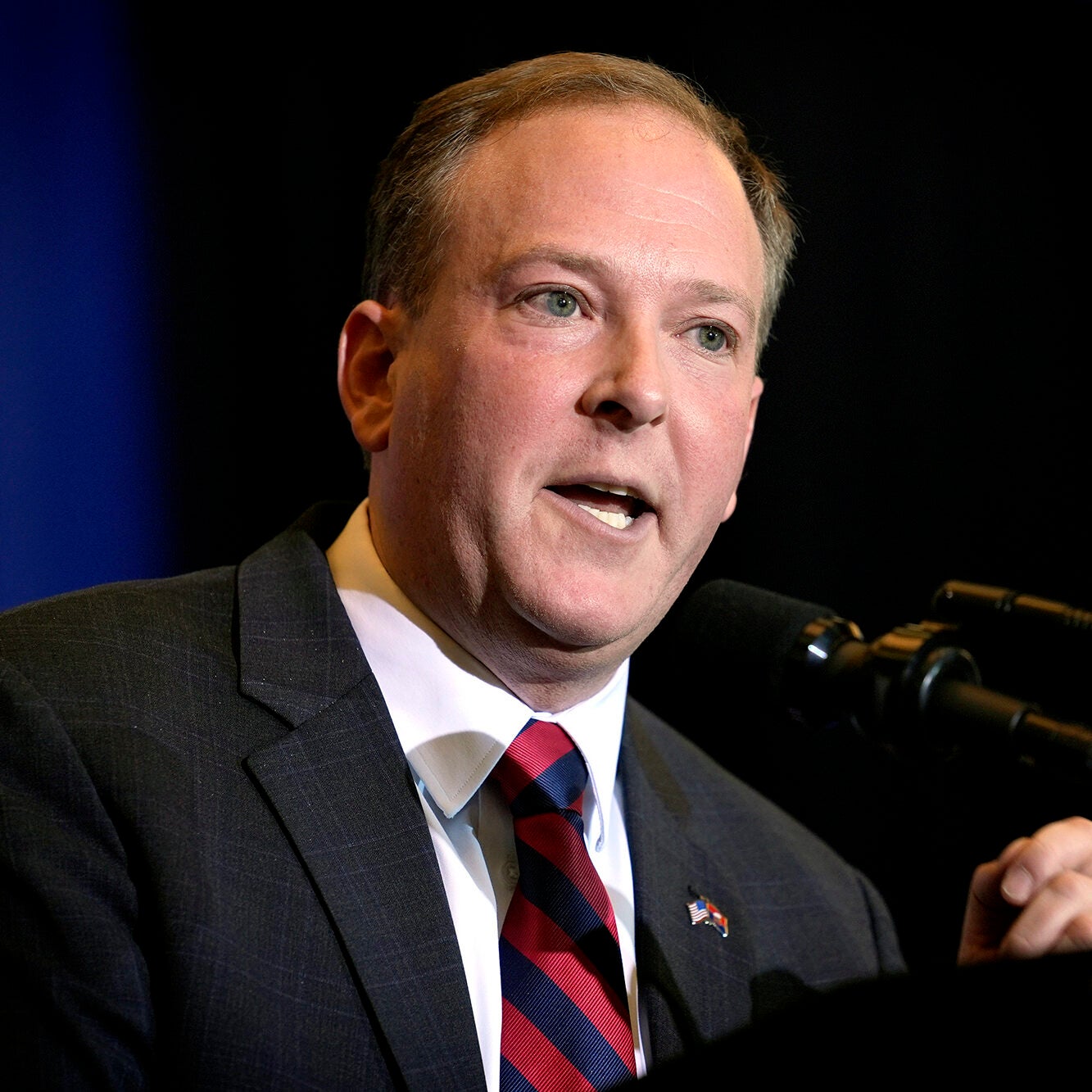 A man speaks at a microphone while wearing a suit with a dark blue and black background.