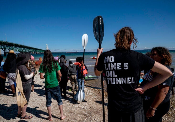 A group of people stand on the bank of a large body of water facing away from the camera, with a large bridge visible on the left of the frame. The shirt on the person on the right reads "NO LINE 5 OIL TUNNEL"