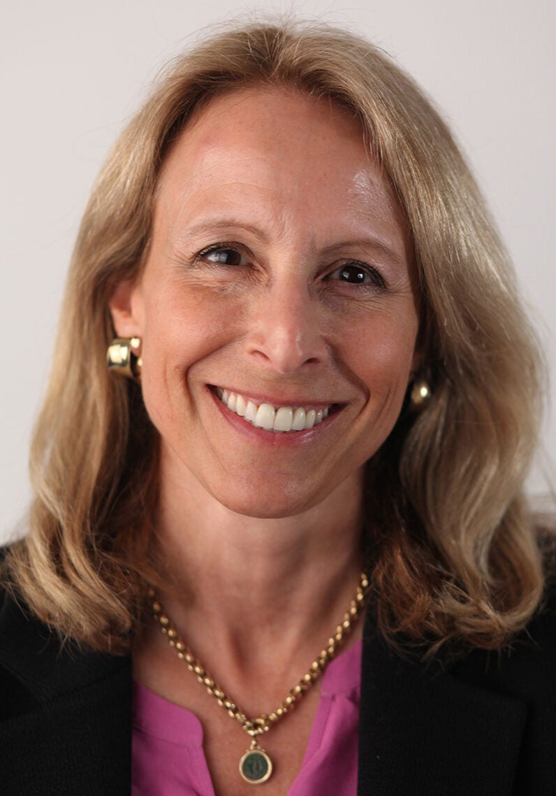 Lisa looks directly at the viewer with a friendly, open smile. She's wearing a dusky pink top under a black blazer, and is in front of a solid beige color background.