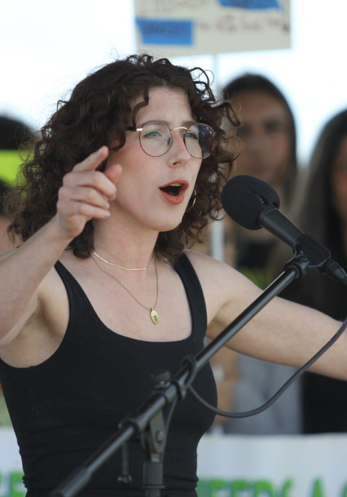 Woman speaking into a microphone with arms raised at a rally.