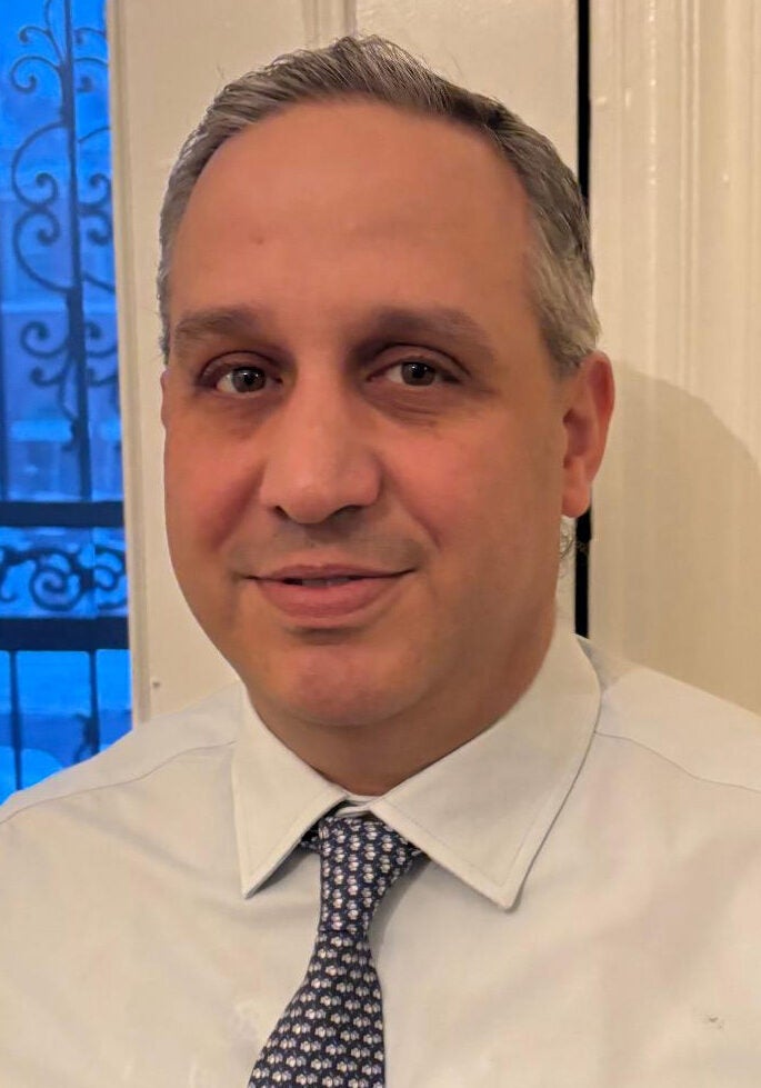 Matthew faces the viewer with a friendly, slight smile. He's wearing a white collar shirt, with a dark patterned tie. Matthew is standing inside a room, in front of a white wall and open window. Visible in the window is a black ornate metal frame, in shadow against the deep blue light of the outdoors.