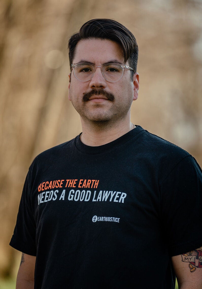 Mychal stands facing the viewer with a somber expression. He's wearing clear framed glasses and a black T-shirt with the text "Because the earth" (in red) "needs a good lawyer" (in white) printed across the front, with the logo of Earthjustice (an icon of the globe on a pedestal, next to the words "Earthjustice). Mychal is standing in an outdoor location. The background is a blurred light brown color.