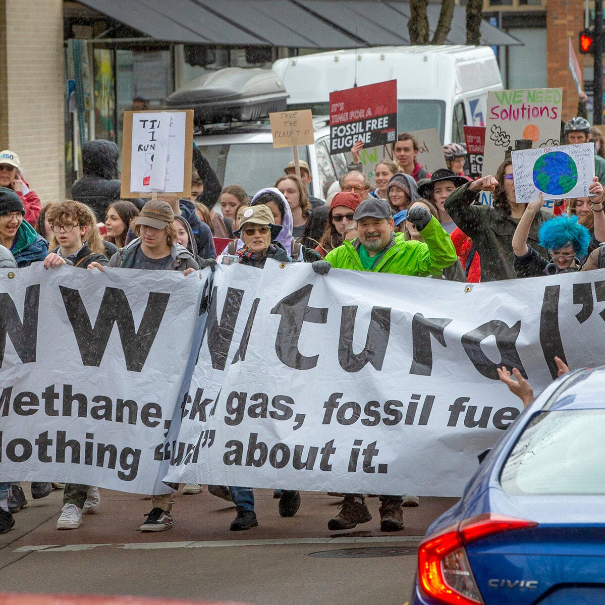 A large group of people, mostly young, march down a street in the rain holding signs against fossil fuels.