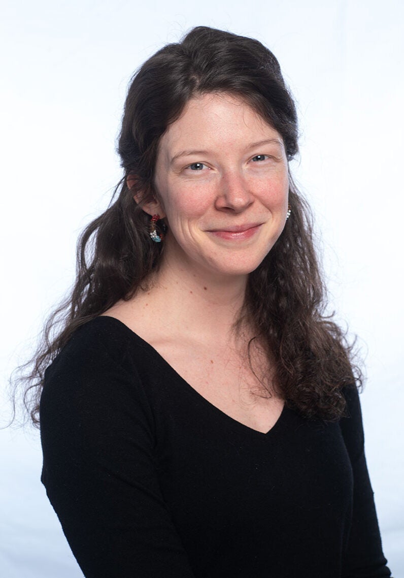 Olivia looks directly at the viewer with a friendly smile. She's wearing a black, long-sleeved shirt and stands in front of a white background. The photo is from the waist up.