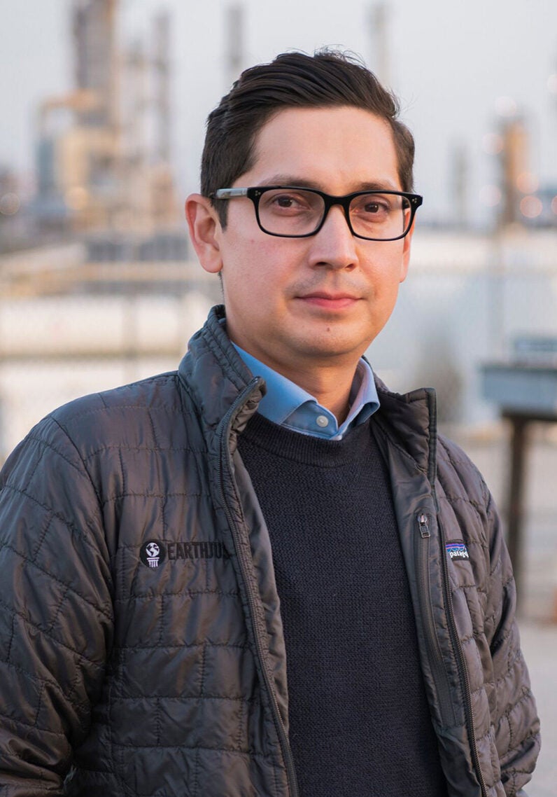 Oscar looks directly at the viewer with a serious expression. He's standing outdoors, in front of an industrial facility, which is partially visible in the blurred background. Oscar is wearing a grey jacket over a black sweater and blue collared shirt. It appears to be an overcast day.