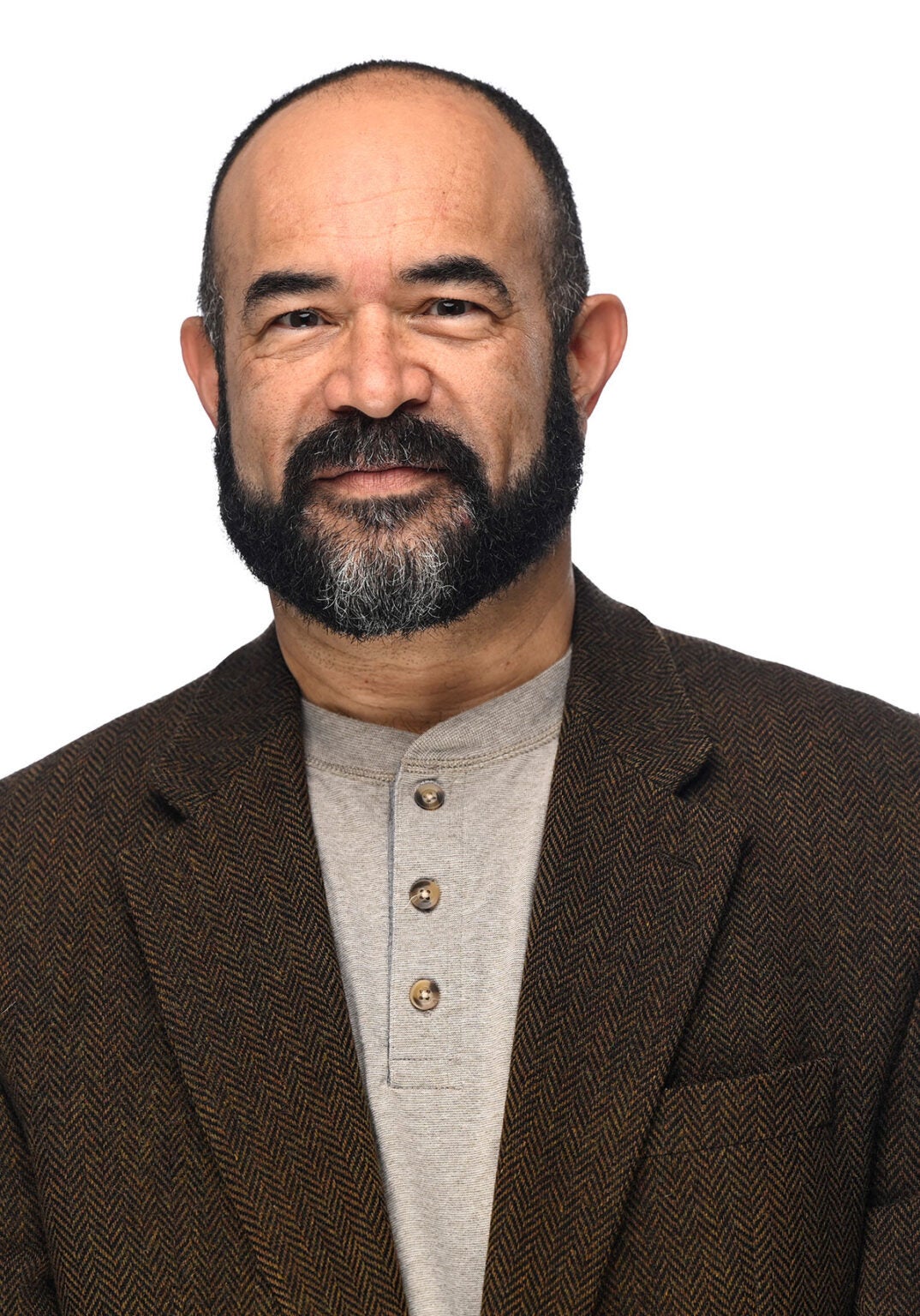 Patrice stands looking directly at the viewer with a slight smile. He's wearing a dark brown suit jacket with a chevron ripple pattern and a light brown knit shirt with a band color. He's standing in front of a white backdrop.
