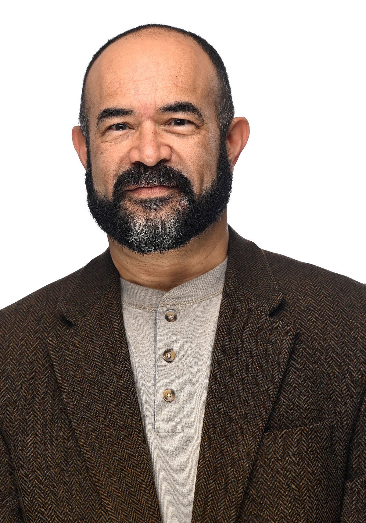 Patrice stands looking directly at the viewer with a slight smile. He's wearing a dark brown suit jacket with a chevron ripple pattern and a light brown knit shirt with a band color. He's standing in front of a white backdrop.