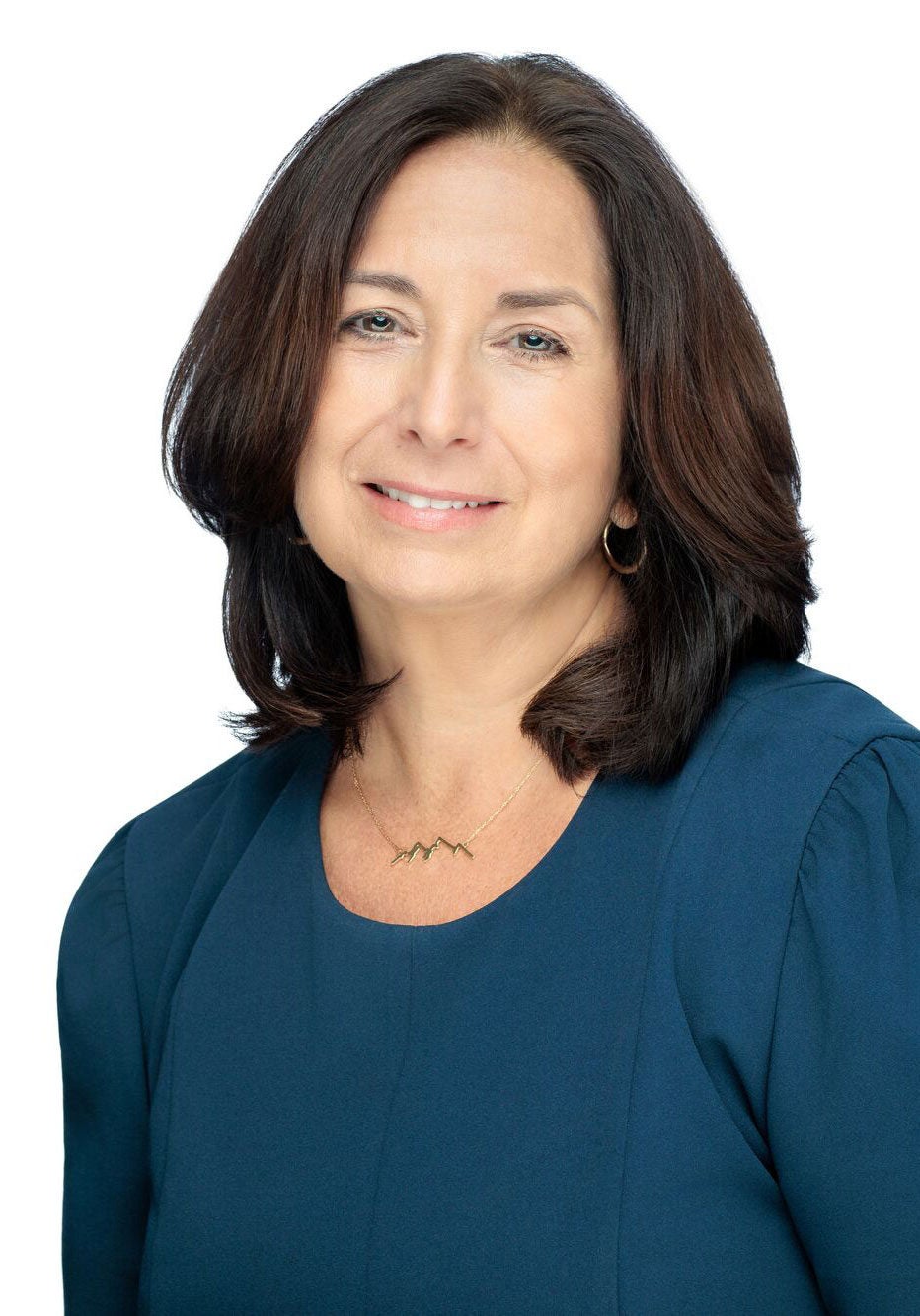 Patricia looks directly at the viewer with a soft smile and kind eyes. Her head is slightly tilted to the viewer's right. She's wearing a dark blue dress with full sleeves. She's standing in front of a white backdrop.