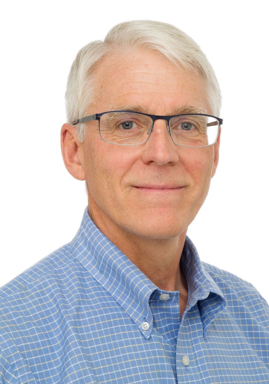 Peter looks directly at the viewer with a slight smile. He's wearing a light blue collared shirt with a thin white vertical and horizontal stripes. He's standing in front of a white backdrop.