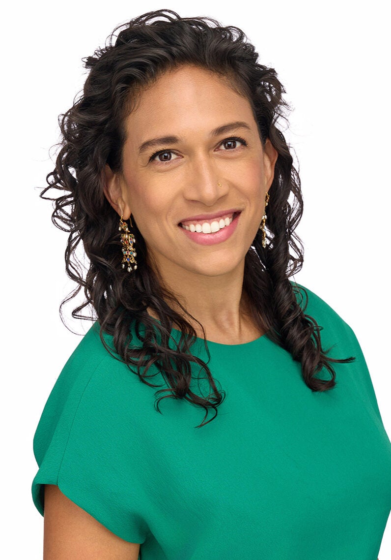 Purba is looking directly at the viewer with a friendly, open smile. Her body is angled slightly to the viewer's right. She's wearing a turquoise top and standing in front of a white backdrop.