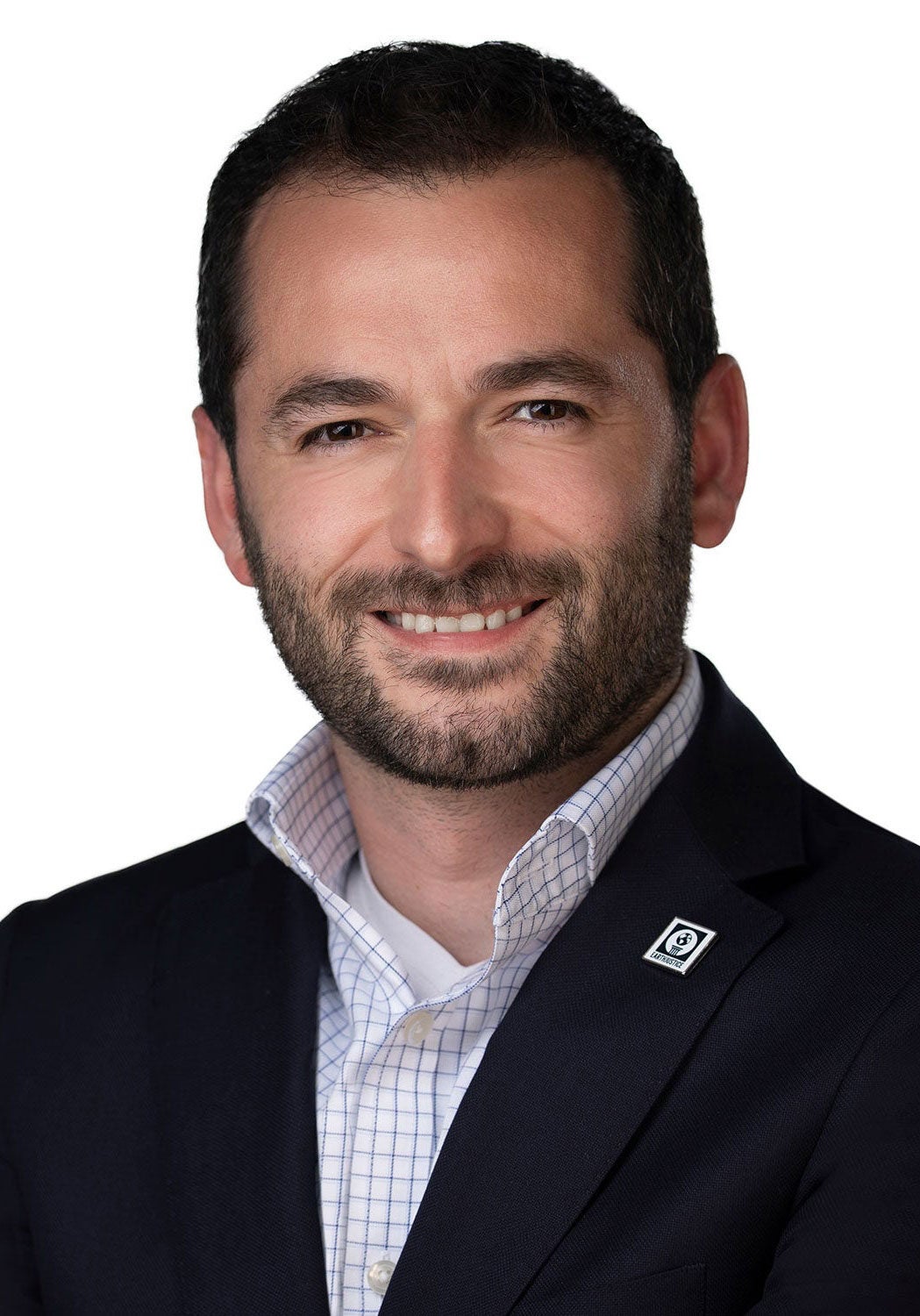 Raúl looks directly at the viewer with a friendly, open smile. He's wearing a black suit jacket and a white collared shirt with thin dark blue vertical and horizontal strips. A square metal pin with Earthjustice's globe logo is pinned to his left suit jacket lapel. He's standing in front of a white backdrop.