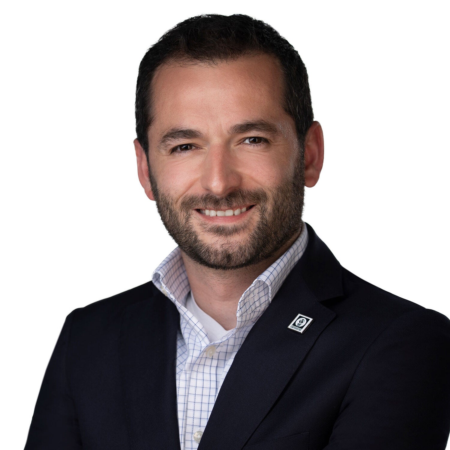 Raúl looks directly at the viewer with a friendly, open smile. He's wearing a black suit jacket and a white collared shirt with thin dark blue vertical and horizontal strips. A square metal pin with Earthjustice's globe logo is pinned to his left suit jacket lapel. He's standing in front of a white backdrop.