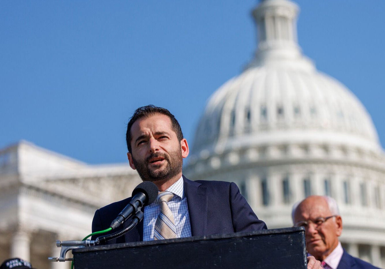 Raúl García, waring a blue suit, speaks into a microphone behind a podium, in front of the U.S. Capitol building under a cloudless blue sky. Several people stand behind him.