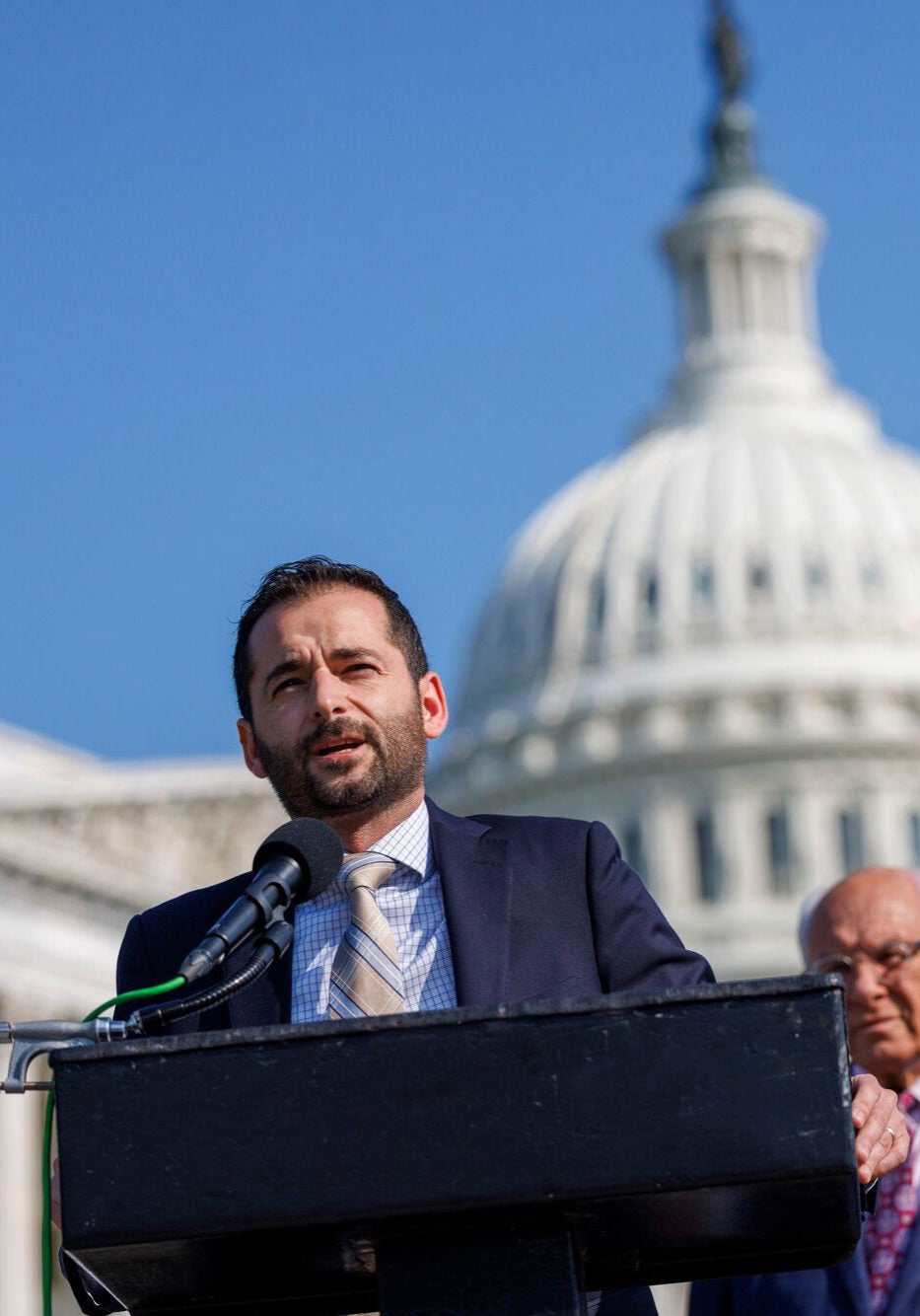 Raúl García, waring a blue suit, speaks into a microphone behind a podium, in front of the U.S. Capitol building under a cloudless blue sky. Several people stand behind him.