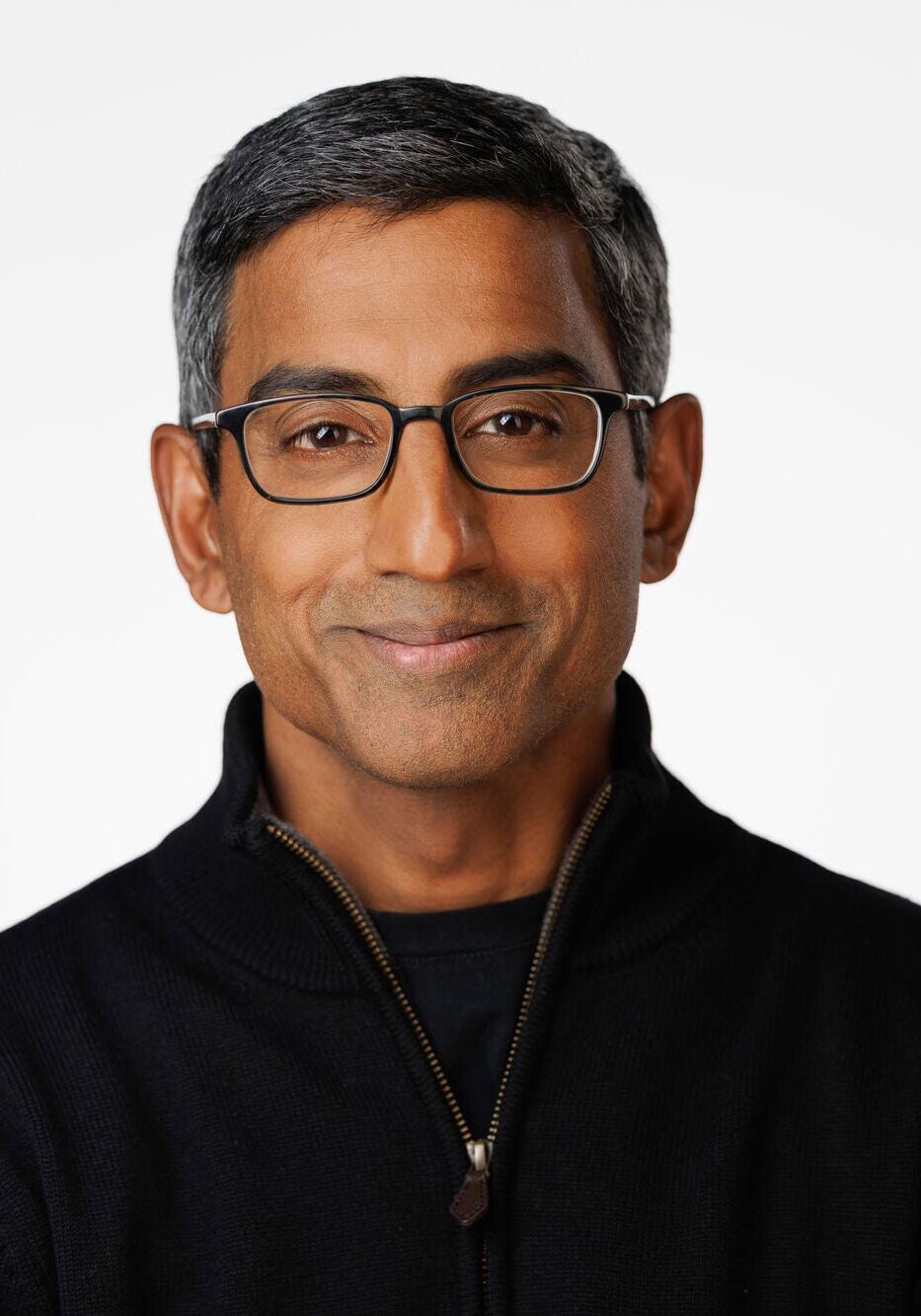 Sam looks directly at the viewer with a slight smile and friendly eyes. He's wearing a black jacket, with the zipper slightly pulled out, and a black shirt. He's standing in front of a white backdrop.