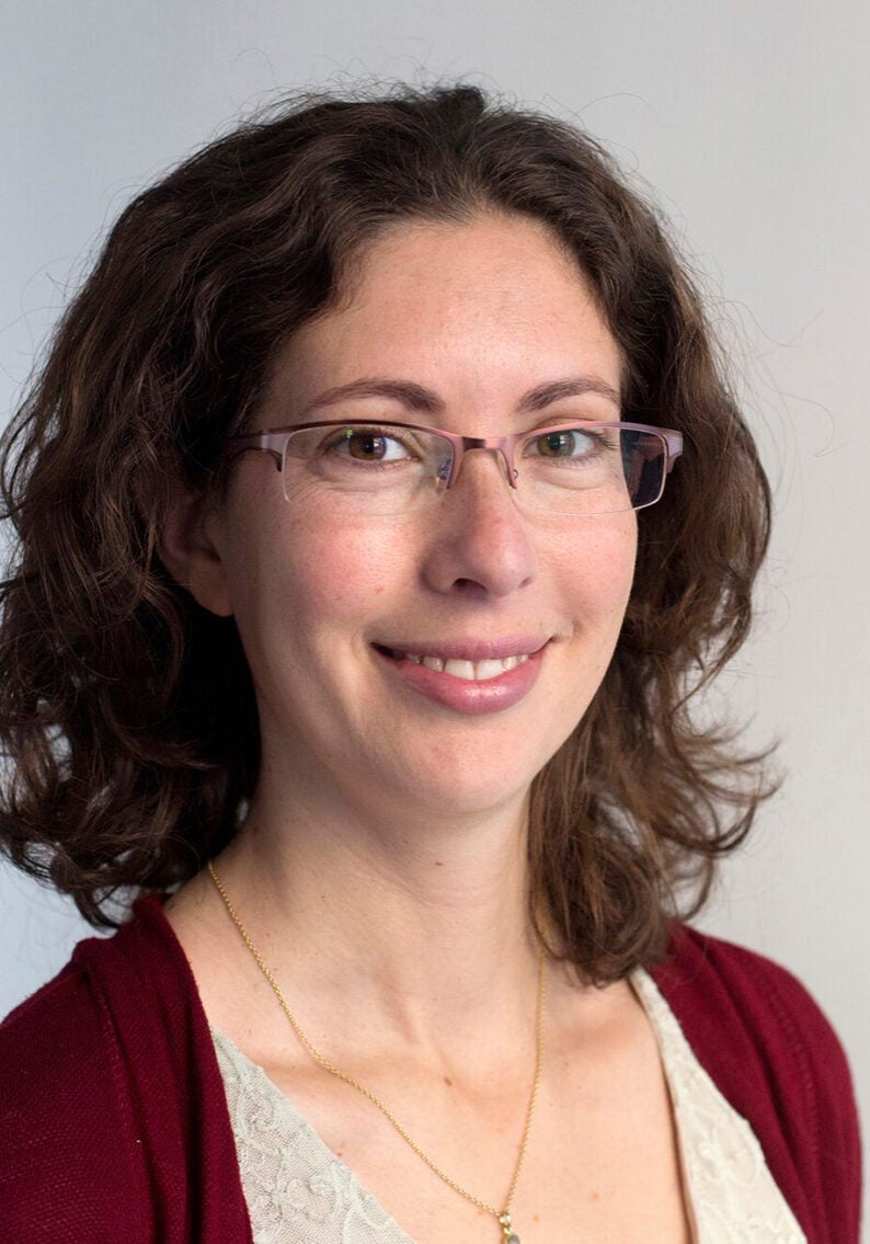 Sara looks directly at the viewer with a friendly, open smile. She's wearing an off-white blouse under a dark red cardigan. She's standing in front of a light-grey backdrop.