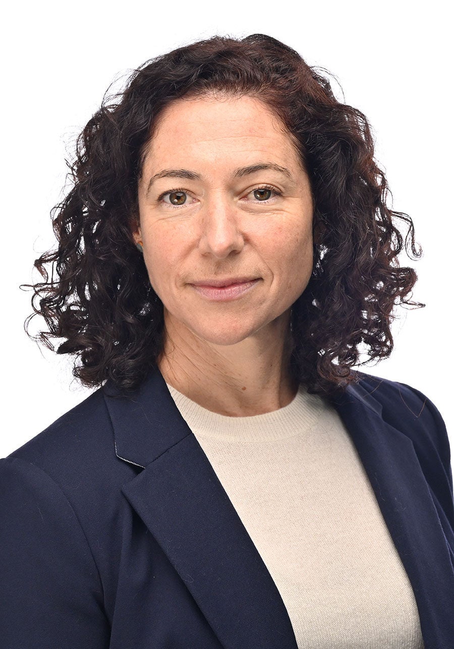 Sarah looks directly at the viewer with a serious expression. She's wearing a dark blue suit jacket and cream knit top. She's standing in front of a white backdrop. Her body is slightly angled to the right of the viewer.
