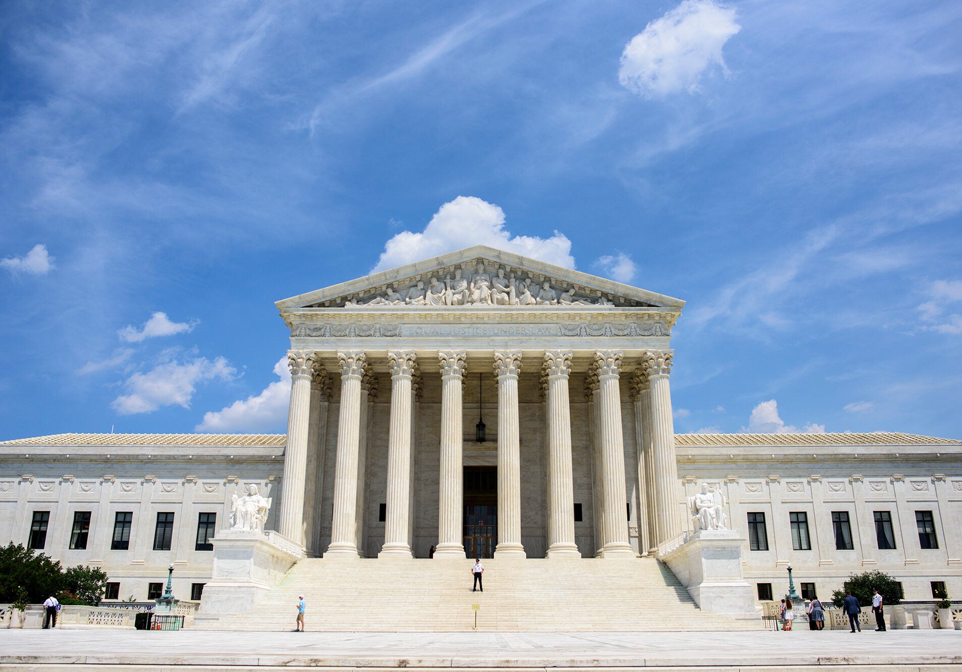 U.S. Supreme Court The U.S. Supreme Court in Washington, D.C., on a sunny day, framed against a blue sky, with a few visitors and security guards standing in front of the building.