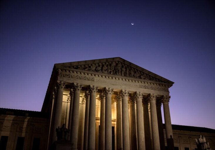 The front of the U.S. Supreme Court building, with its entrance brightly lit against a dark purple sky.