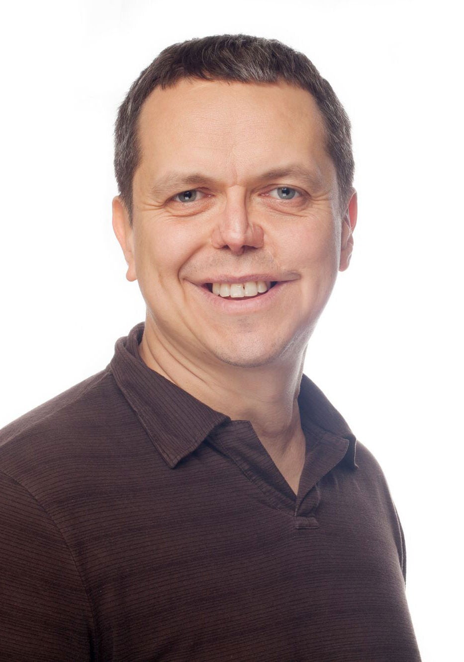 Shannon looks directly at the viewer with a friendly, open smile. He's wearing a brown polo shirt with thin, darker brown horizontal striped pattern of varied widths. He's standing in front of a white backdrop.