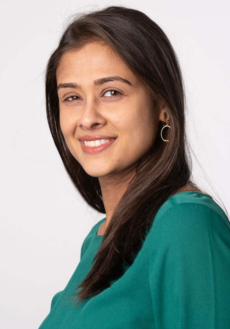 Sharmeen looks directly at the viewer with a friendly, open smile. Her body is angled to the viewer's left. Sharmeen is wearing a green, long-sleeved top and is standing in front of an off-white backdrop.
