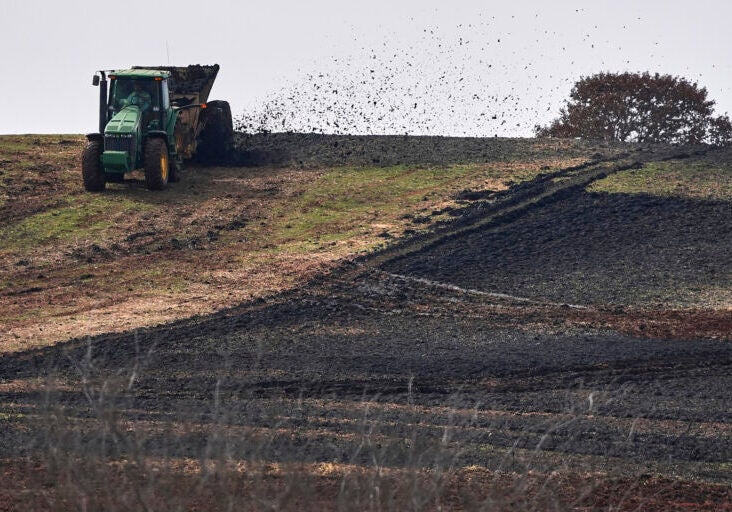 A tractor blows dark, Earth-like material out of a trailer and onto a tilled field.