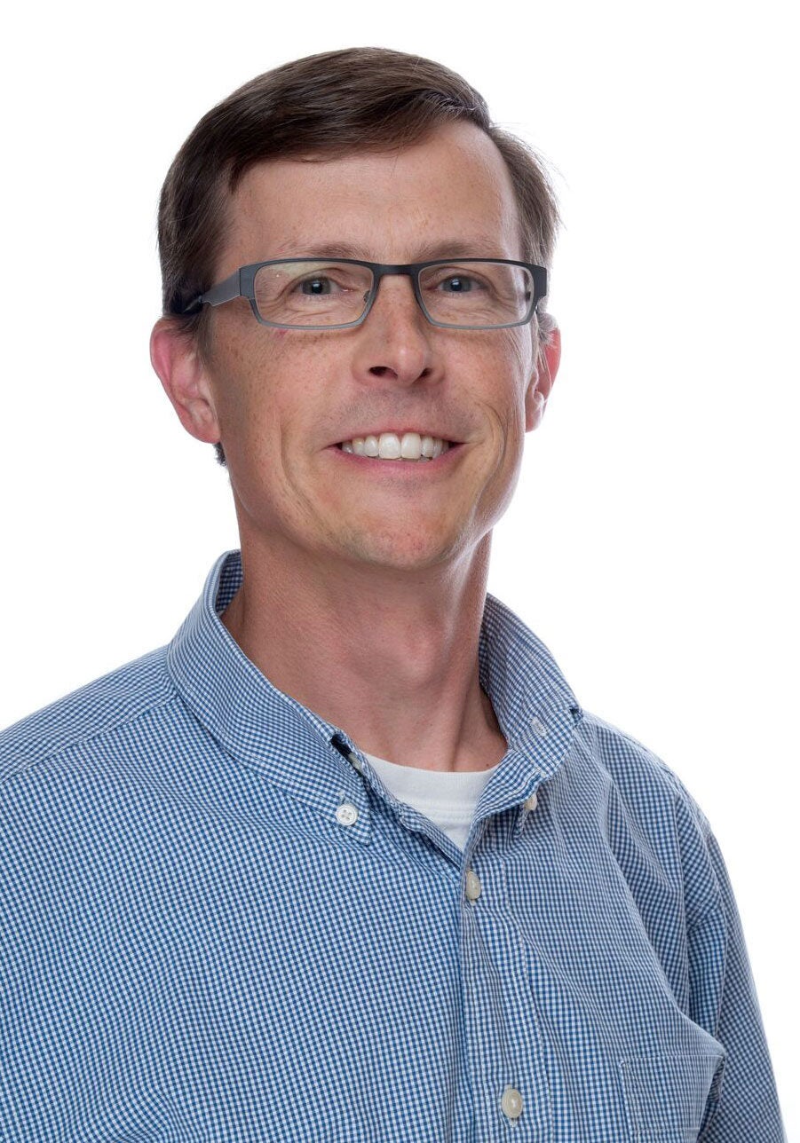 Steve looks directly at the viewer with a cheerful, open smile. He's wearing a collared button-down shirt with a white and blue checkered pattern. He's standing in front of a white backdrop.