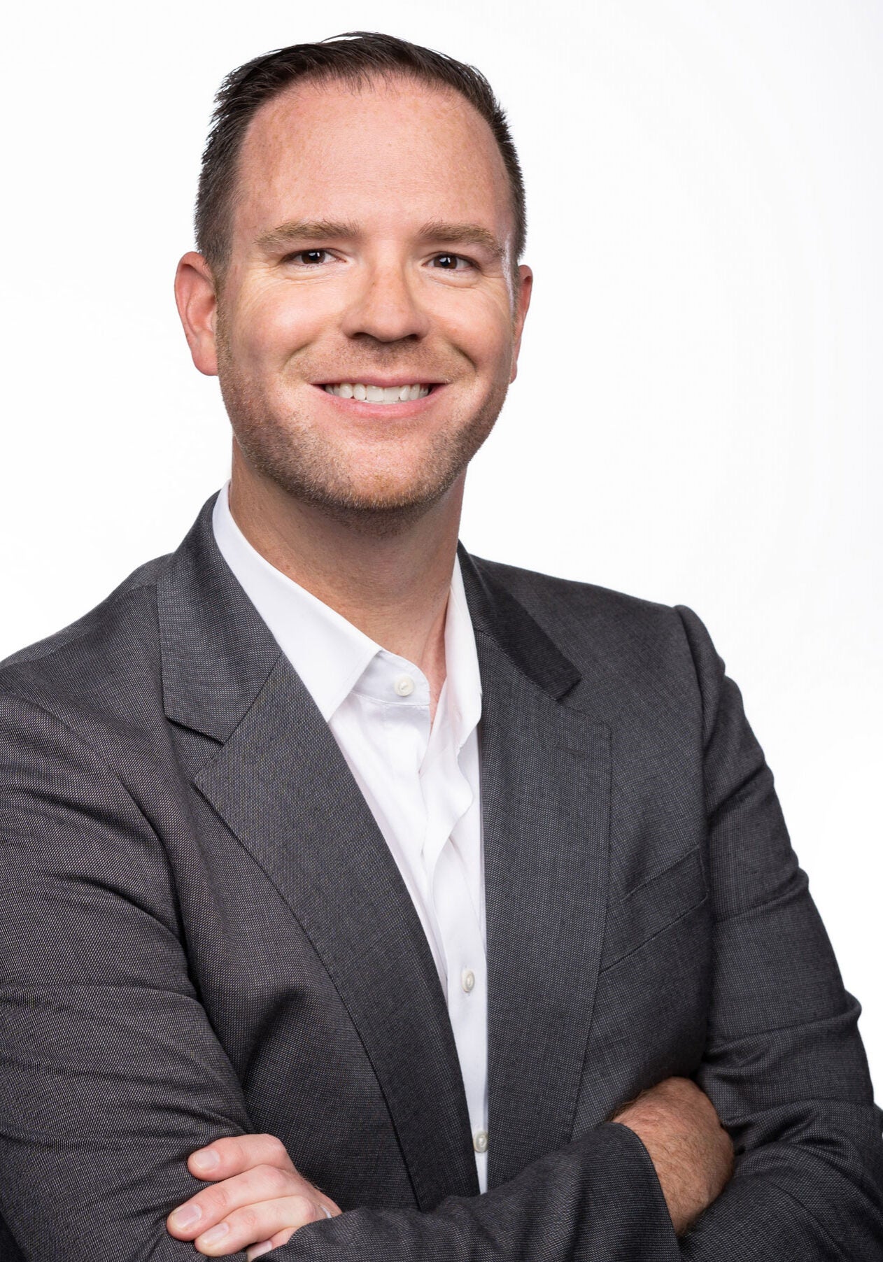 Steve looks directly at the viewer with a friendly, open smile. His arms are crossed in a relaxed way across his chest. He's wearing a dark grey suit with a white collared shirt, standing in front of a white backdrop.