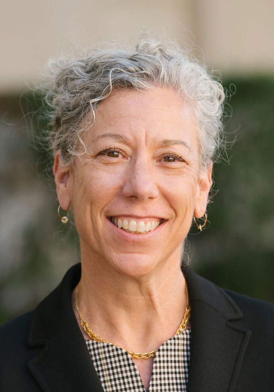 Susan looks directly at the viewer with an open, friendly smile. She's wearing a black suit jacket and a black and white checkered blouse. She's standing outdoors. The background is blurred, with tan and dark green colors.