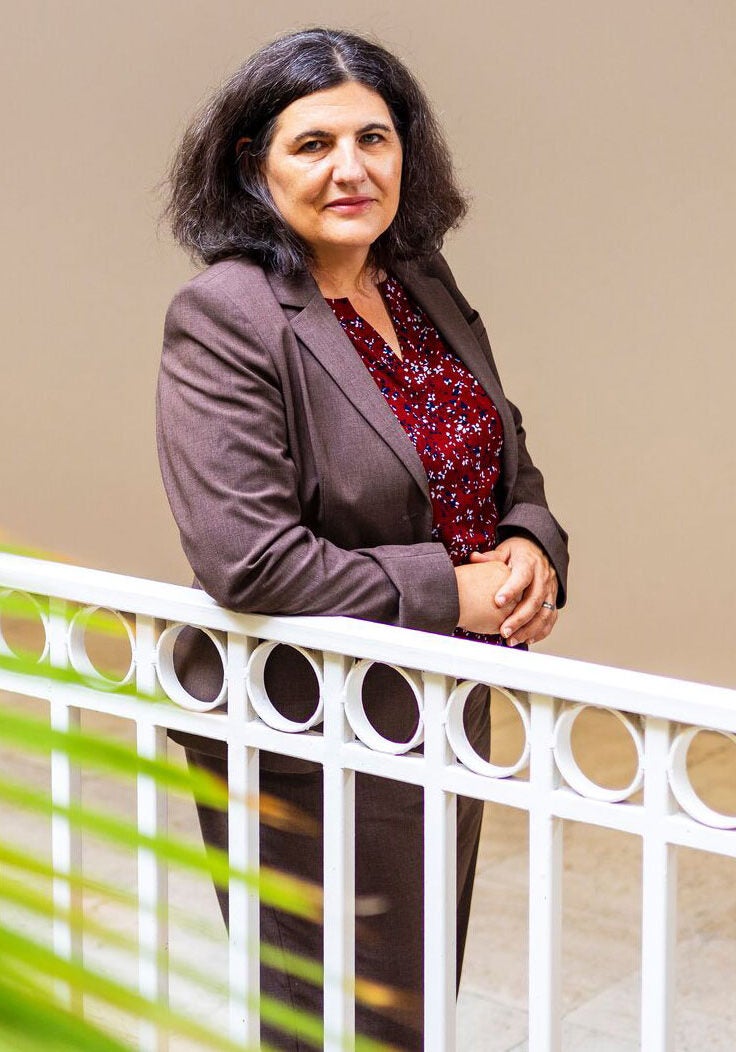 Tania, wearing a burgundy suit and dark red patterned blouse, leans against a white metal railing, looking directly at the viewer with a serious, faint smile. The edges of a palm frond are blurred, in the foreground's bottom left corner of the image.