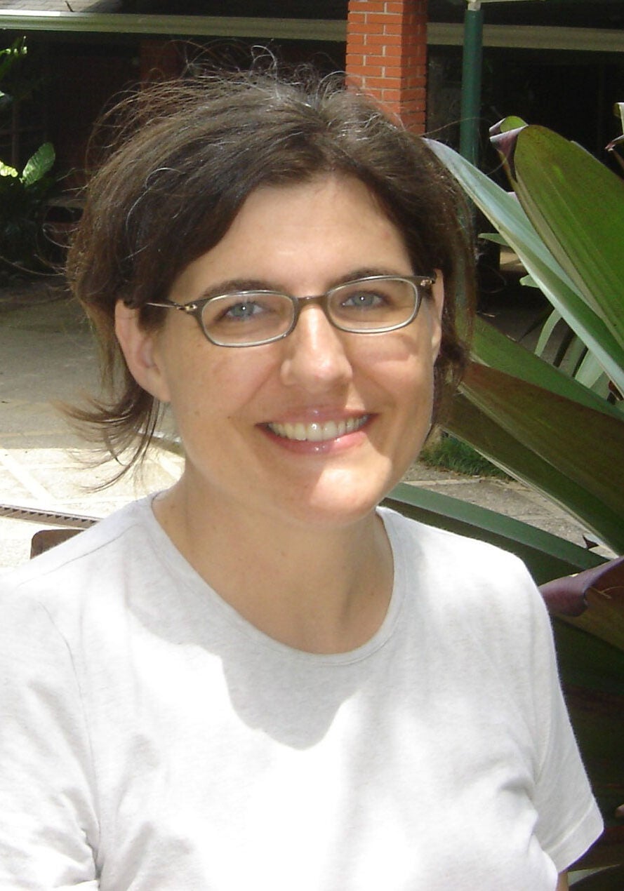 Tania looks directly at the viewer with a friendly, open smile. She's wearing a white t-shirt in a outdoor setting. A large agave plant is partially visible behind her.