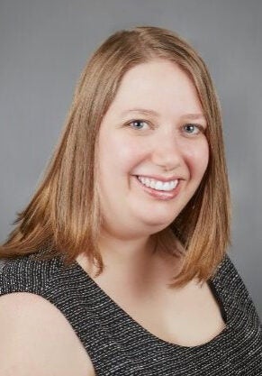 Torie looks at the viewer with a friendly, open smile. Her body is angled towards the viewer's right. She's wearing a black and white speckled sleeveless top and is in front of a grey backdrop.